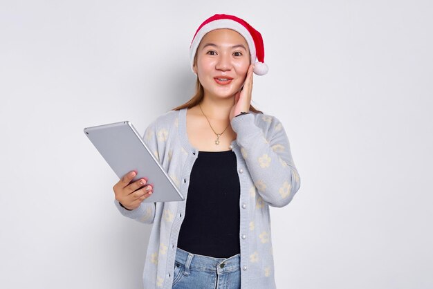 Excited young Asian woman in a hat Christmas using a digital tablet and touching her cheek isolated over white background Indonesian people celebrate Christmas concept