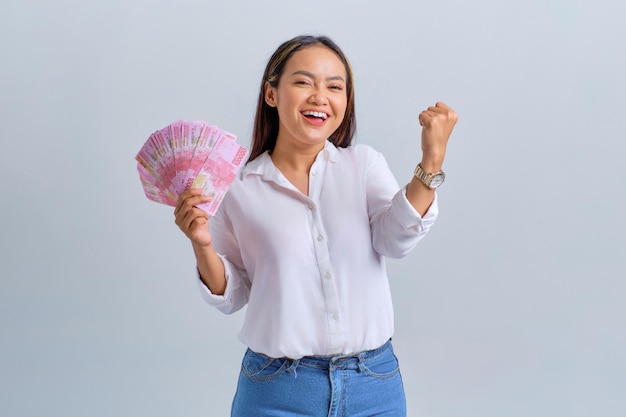 Photo excited young asian woman holding money banknotes and making success gestures isolated over white background