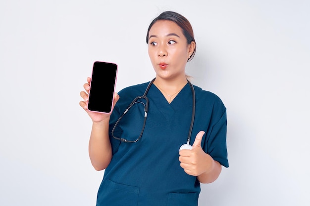 Возбужденная молодая азиатская медсестра, работающая в синей форме, показывает мобильный телефон с пустым экраном и поднимает большой палец вверх на белом фоне