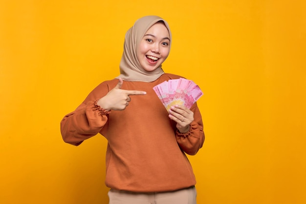 Excited young Asian woman in orange shirt points at money banknotes isolated over yellow background