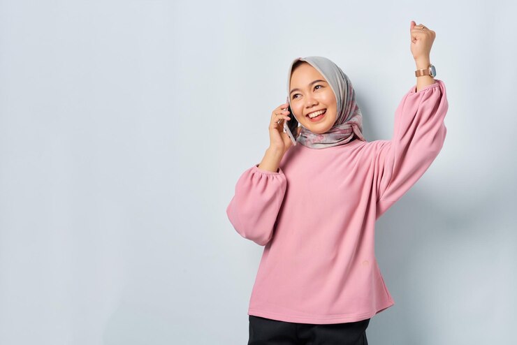 Excited young Asian woman in pink shirt talking on mobile phone and doing winner gesture isolated over white background