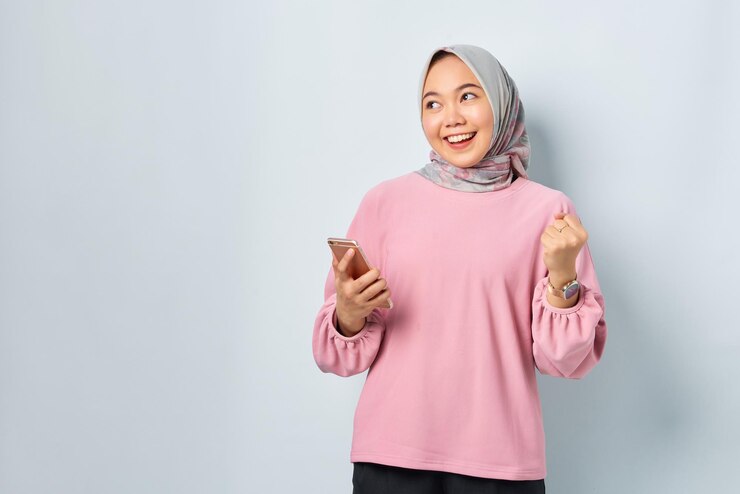 Excited young Asian woman in pink shirt using mobile phone and doing winner gesture looking away at blank space isolated over white background
