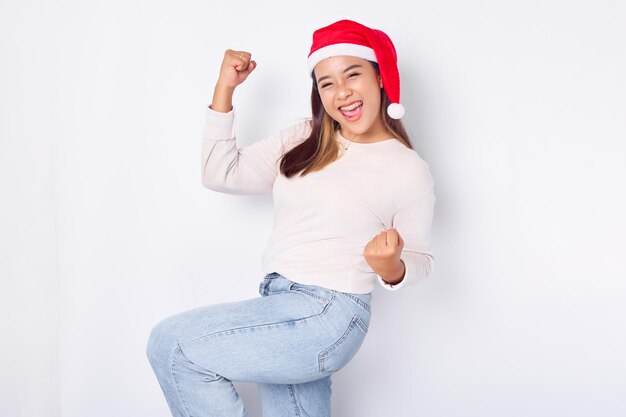 Excited young Asian woman in Santa Claus hat celebrating success with raised fists isolated over white background celebration Christmas holiday and New Year concept