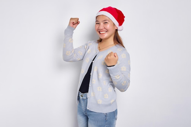 Excited young Asian woman in Santa Claus hat celebrating success with raised fists isolated over white background