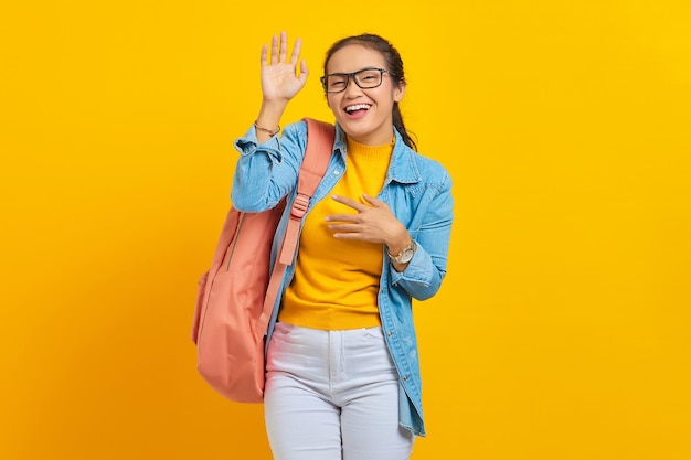 黄色の背景にカメラに手を振っているバックパックとデニムの衣装で興奮した若いアジアの女性学生大学の大学の概念の教育