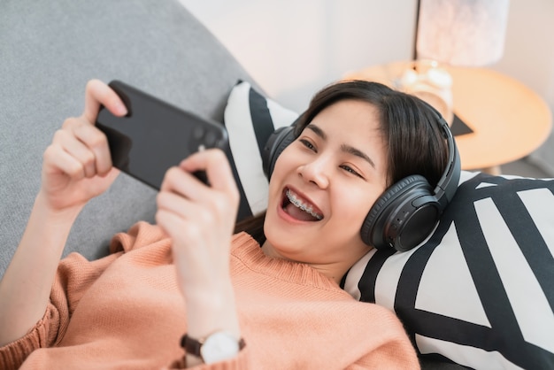 Excited young Asian woman wears white headphones and playing games on smartphone in the living room at home.