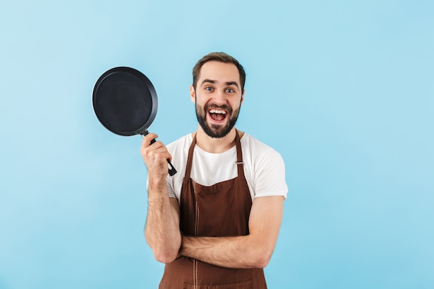 Excited young bearded man cook wearing apron standing isolated over blue wall, showing frying pan