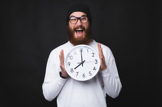 An excited young and bearded man is holding a big white clock on black background.