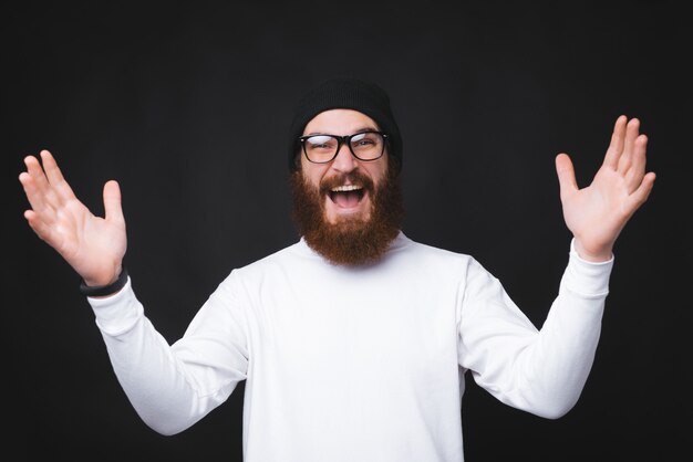 Excited young and bearded man is holding both of his hands up on black wall.