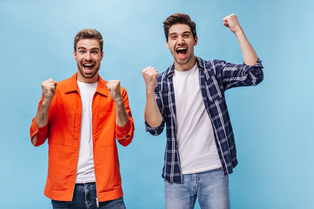 Excited young bearded man in orange jacket and his friend in checkered shirt rejoice and smile on blue wall.