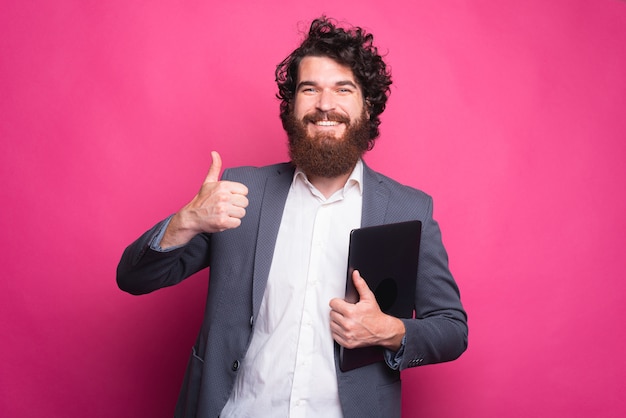 Excited young bearded man in suit showing thumb up and holding laptop