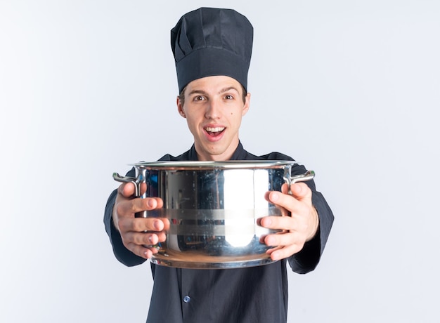 Excited young blonde male cook in chef uniform and cap stretching out pot towards camera looking at camera isolated on white wall