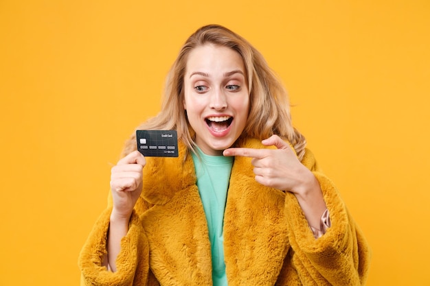 Photo excited young blonde woman girl in yellow fur coat posing isolated on orange background studio portrait. people emotions lifestyle concept. mock up copy space. point index finger on credit bank card.