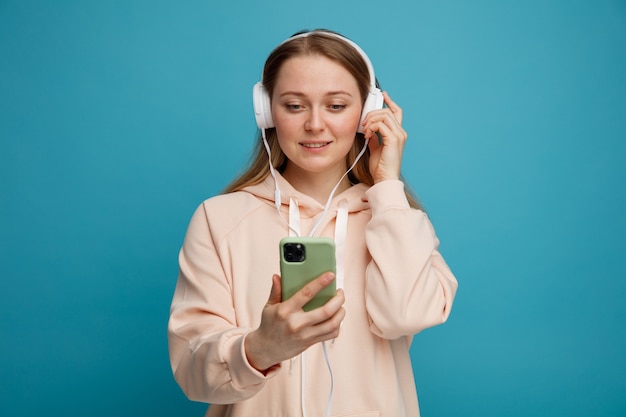 Excited young blonde woman wearing and grabbing headphones holding and looking at mobile phone 