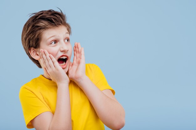 Excited young  boy look aneway and screaming in yellow t-shirt isolated on blue wall, copy space