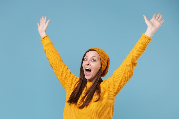 Excited young brunette woman girl in yellow sweater and hat\
posing isolated on blue background studio portrait. people sincere\
emotions lifestyle concept. mock up copy space. rising spreading\
hands.