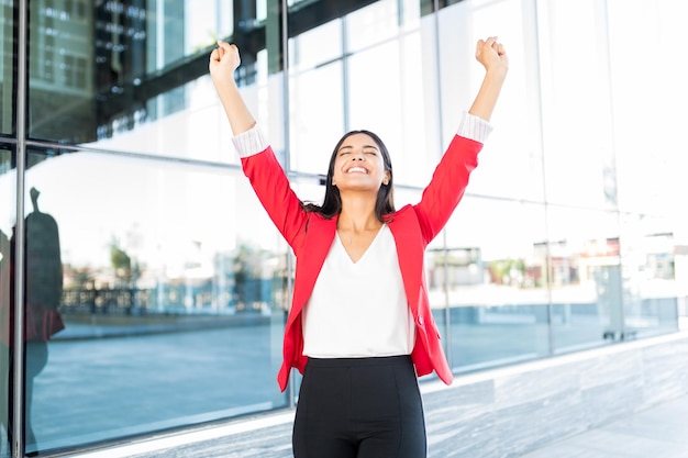 Excited young businesswoman with arms raised standing outside office building