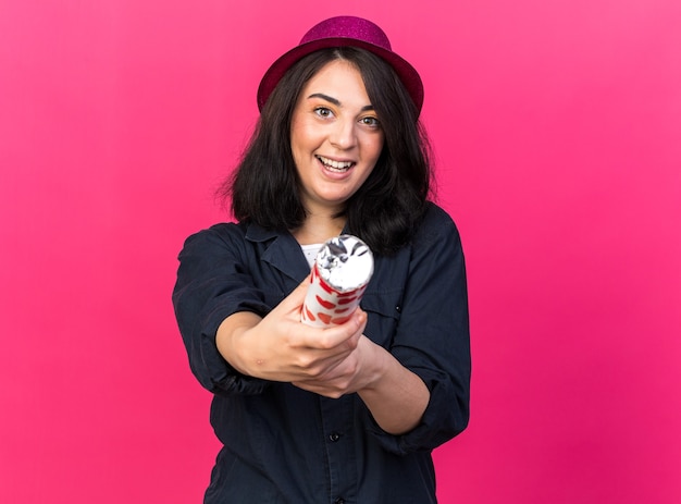 Excited young caucasian party woman wearing party hat pointing at front with confetti cannon looking at front isolated on pink wall