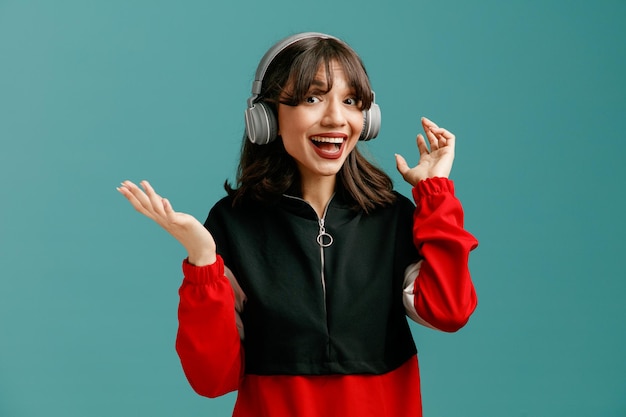 Excited young caucasian woman wearing headphones looking at camera showing empty hands isolated on blue background