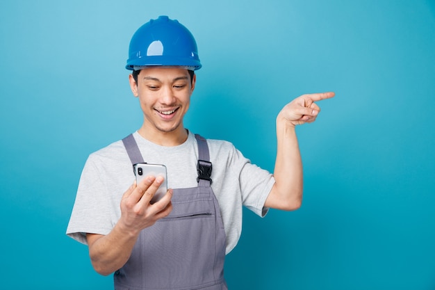 Eccitato giovane operaio edile che indossa il casco di sicurezza e tenuta uniforme e guardando il telefono cellulare che punta a lato