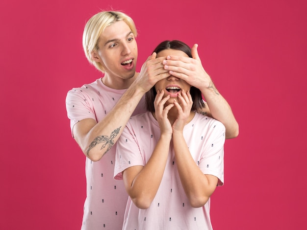 Photo excited young couple wearing pajamas man standing behind woman covering her eyes with hands looking at her woman touching chin with hands isolated on pink wall