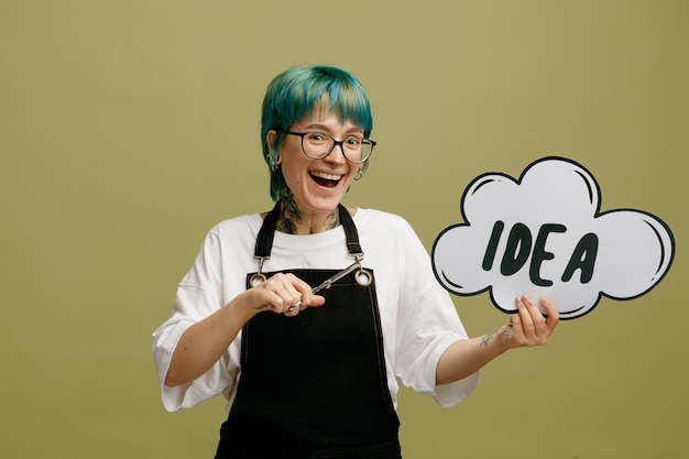 Excited young female barber wearing glasses uniform showing idea bubble looking at camera pointing with scissors at idea bubble isolated on olive green background