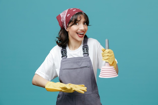 Excited young female cleaner wearing uniform bandana and rubber gloves holding plunger pointing at it with hand looking at camera isolated on blue background