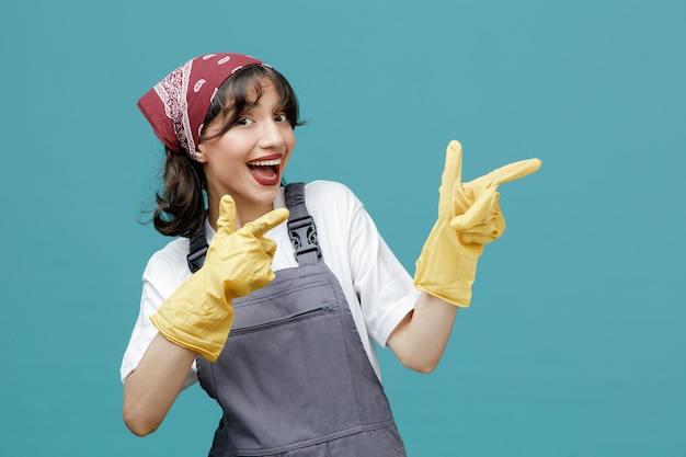 Взволнованная молодая женщина-уборщица в униформе бандана и резиновых перчатках смотрит в камеру, указывая пальцами в сторону, изолированную на синем фоне