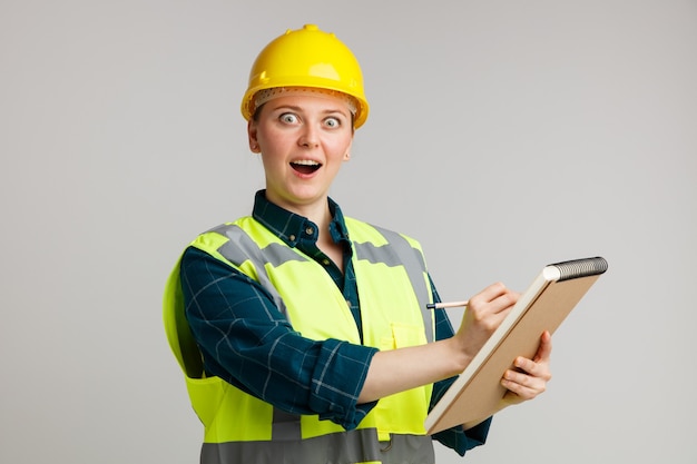 Excited young female construction worker wearing safety helmet and safety vest holding notepad pointing pencil on it 