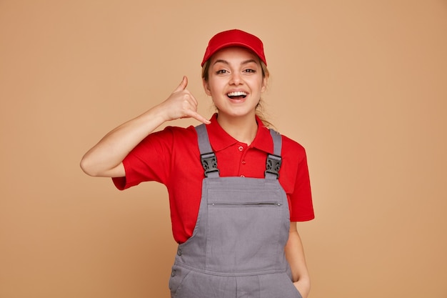 Foto emozionato giovane operaio edile femminile che indossa l'uniforme e berretto tenendo la mano in tasca facendo gesto di chiamata