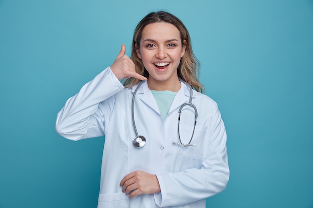 Excited young female doctor wearing medical robe and stethoscope around neck doing call gesture 