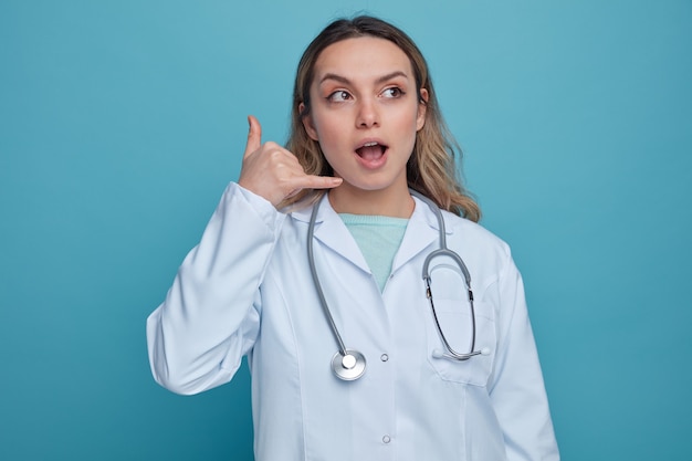 Excited young female doctor wearing medical robe and stethoscope around neck looking at side doing call gesture 