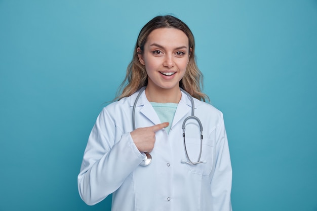 Foto eccitato giovane medico femminile che indossa abito medico e stetoscopio intorno al collo che punta a se stessa