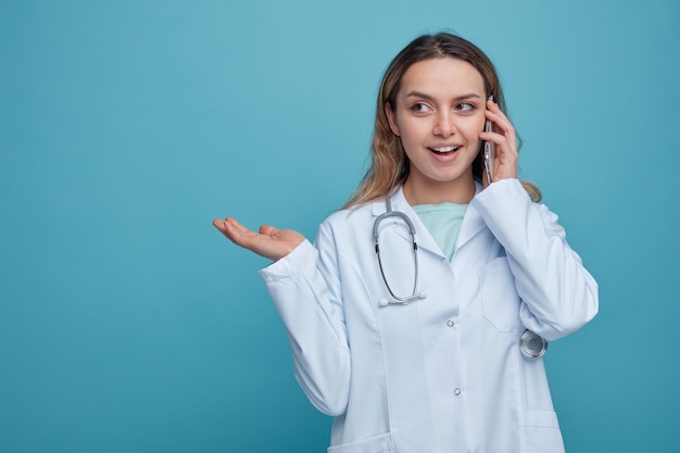 Excited young female doctor wearing medical robe and stethoscope around neck talking by phone looking at side showing empty hand 