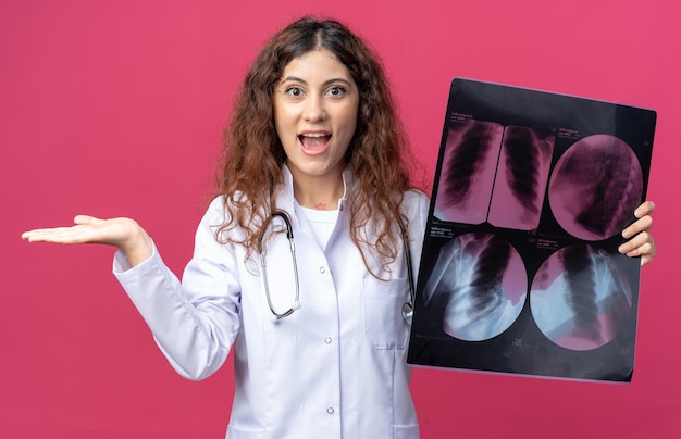 Excited young female doctor wearing medical robe and stethoscope showing x-ray shot to camera looking at front and showing empty hand isolated on pink wall