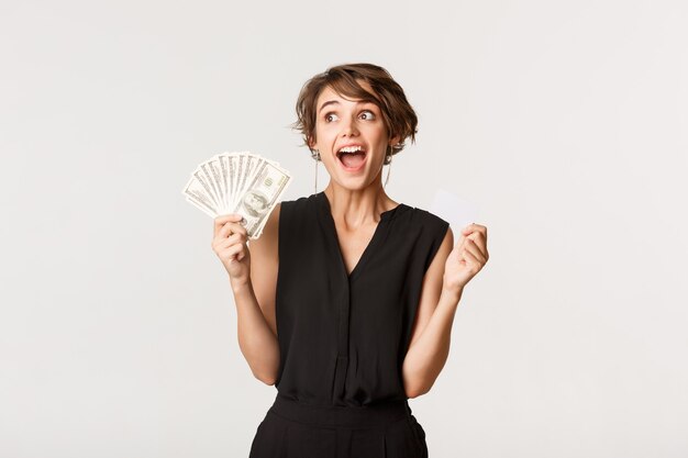 Excited young female shopaholic yelling from thrill and holding credit card with money