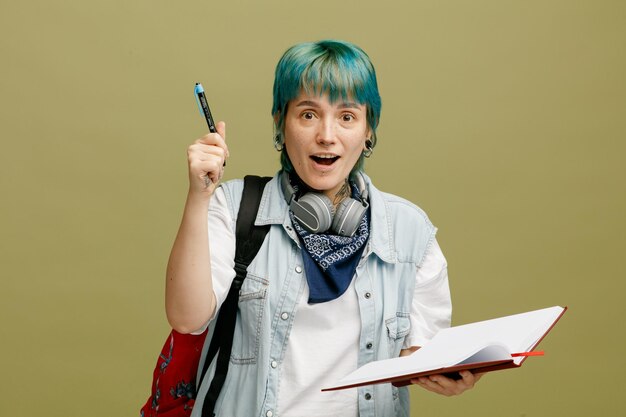 Eccitata giovane studentessa che indossa cuffie e bandana sul collo e zaino con in mano un taccuino aperto guardando la fotocamera rivolta verso l'alto con penna isolata su sfondo verde oliva