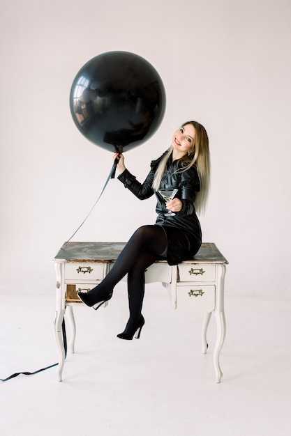 Excited young girl in black dress celebrating holding black balloon and sitting on white vintage table isolated on white background. International Women's Day, Happy New Year, birthday party concept