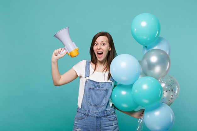 Giovane ragazza eccitata in abiti di jeans che tiene la bocca aperta tenere il megafono che celebra con palloncini colorati isolati su sfondo blu turchese della parete. concetto di emozioni della gente della festa di compleanno.