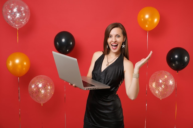 手を広げて、真っ赤な背景の気球で祝っている間、ラップトップPCコンピューターで作業している小さな黒いドレスを着た興奮した少女。明けましておめでとう、誕生日のモックアップホリデーパーティーのコンセプト。