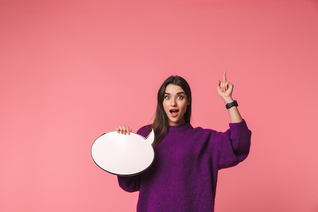 Excited young girl wearing sweater standing isolated over pink, holding empty speech bubble, pointing