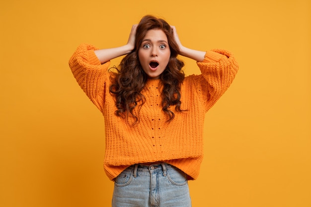 Excited young girl in yellow sweater posing in studio with wavy hair isolated on yellow wall