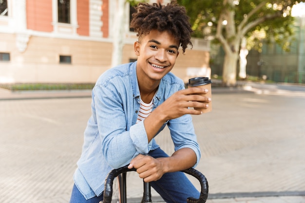 Excited young guy walking outdoors with bicycle holding coffee