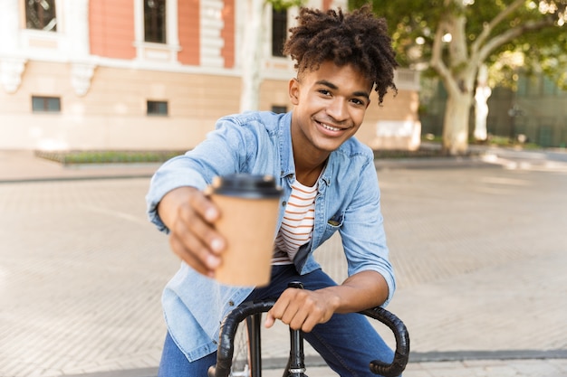 Excited young guy walking outdoors with bicycle holding coffee