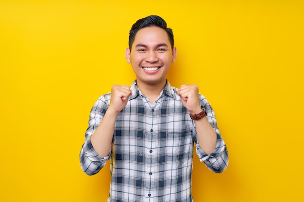 Excited young handsome Asian man celebrating victory or success with raised fists up isolated on yellow background People lifestyle concept