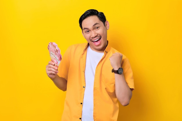 Excited young handsome Asian man holding bunch of money and celebrating success isolated on yellow background