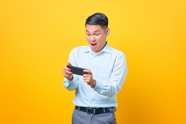 Excited young handsome businessman playing a game on mobile phone on yellow background