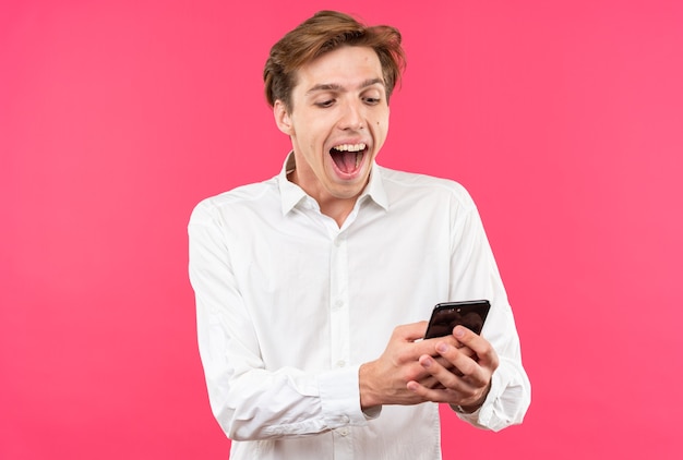 Возбужденный молодой красивый парень в белой рубашке держит и смотрит на телефон, изолированный на розовой стене