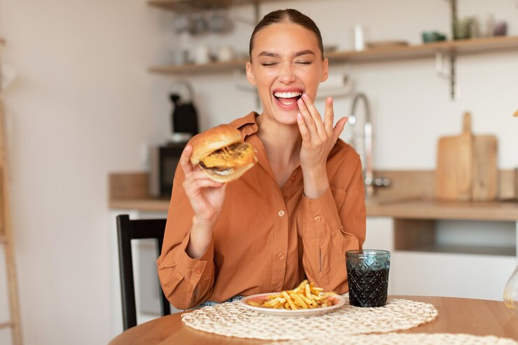 Photo excited young lady eating burger and laughing eating a harmful food sitting at table in kitchen