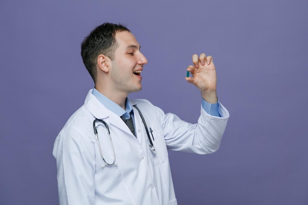 excited young male doctor wearing medical robe and stethoscope around neck standing in profile view holding capsule in front of mouth looking at it with open mouth isolated on purple background